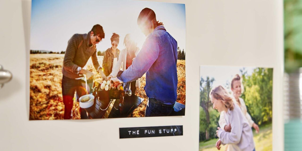 An einen cremefarbenen Kühlschrank hängen zwei Sofortfotos. Das linke Foto zeigt vier junge Menschen beim Grillen auf einem Feld. Auf dem rechten Foto sind zwei lachende Frauen zu sehen, die in einem Garten stehen. Zwischen den Fotos befindet sich ein geprägtes Etikett, auf dem „The fun stuff“ steht.