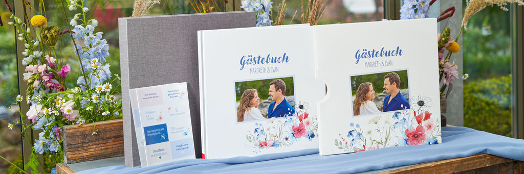 Ein Fotobuch, das als Gästebuch für eine Hochzeit gestaltet wurde, steht auf einem Tisch. Daneben steht der Schuber im gleichen Design. Auf dem Cover ist ein Foto des Brautpaares zu sehen. Neben dem Buch stehen Fotosticker-Bögen. Im Hintergrund befindet sich Blumendeko.