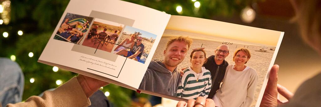 Blick über die Schulter: Papa Olaf und Mama Birgit halten das CEWE FOTOBUCH in ihren Händen. Auf der aufgeschlagenen Doppelseite sind die beiden mit ihren Kindern am Strand zu sehen. Im Hintergrund funkeln die Lichter des Weihnachtsbaumes.