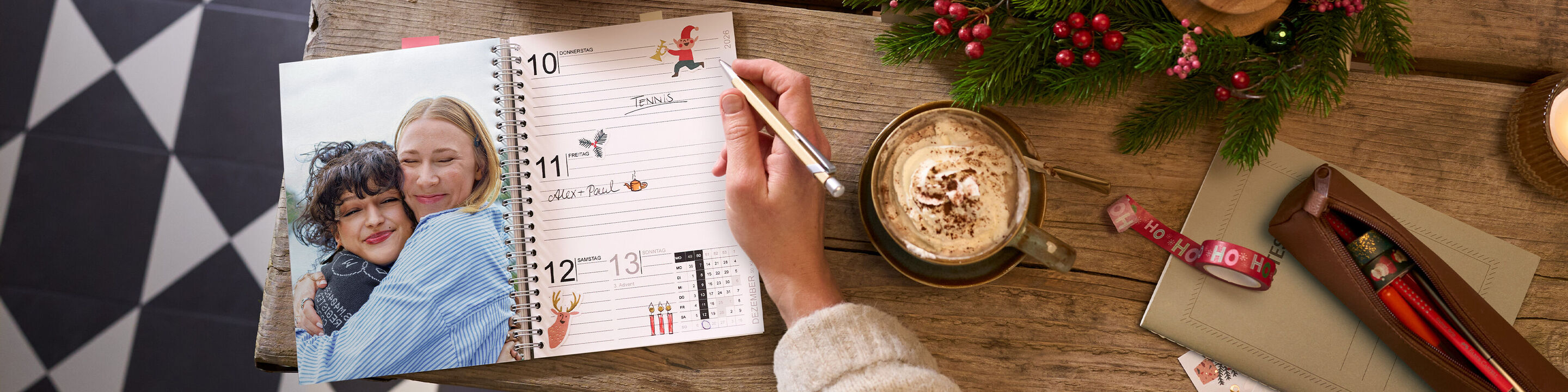 Offener CEWE Fotokalender mit Terminseite und großem Foto von zwei Freundinnen auf einem Holztisch neben Tasse und Deko