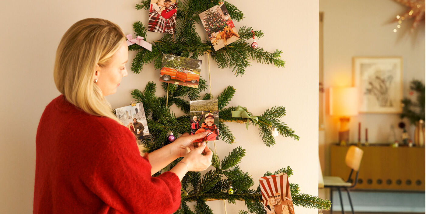 Person hängt liebevoll Fotos an einen Tannenbaum aus Zweigen an der Wand und gestaltet so eine persönliche Fotodeko