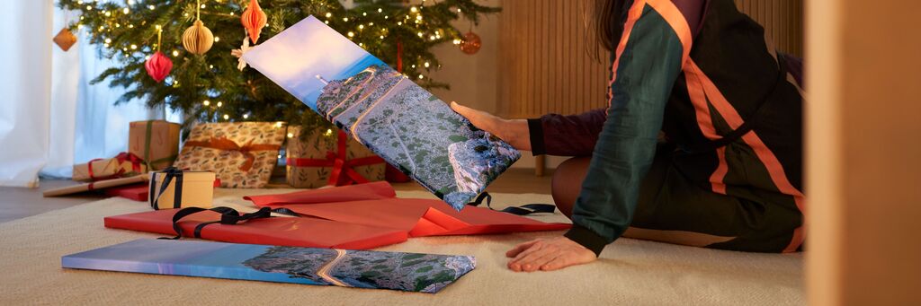 Das Foto zeigt ein Wohnzimmer mit festlich dekoriertem Tannenbaum. Davor sitzt eine Frau neben Geschenken auf dem Boden. In einer Hand hält sie den Teil eines Wandbildes, auf dem ein Landschafts-Motiv abgebildet ist, ein weiterer liegt vor ihr.