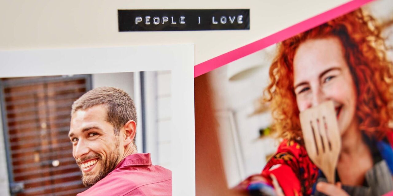 An einem cremefarbenen Kühlschrank hängen zwei Sofortfotos. Links ist ein junger Mann, rechts eine lachende Frau zu sehen, die sich einen Pfannenwender vor das Gesicht hält. Über den Fotos befindet sich ein mit Text geprägtes Etikett, auf dem „People I Love“ steht.