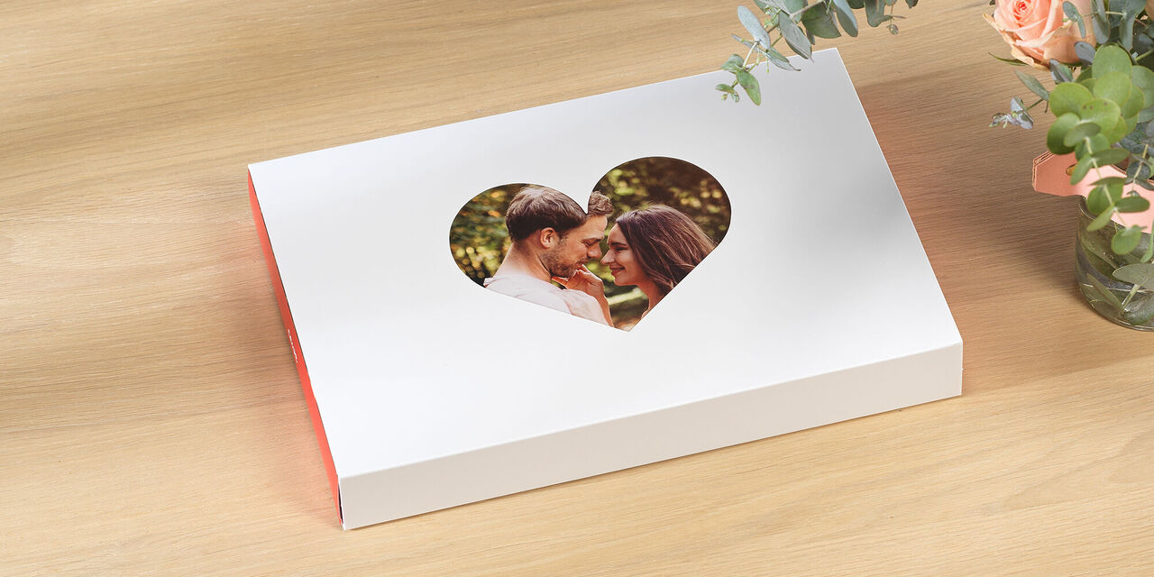 Geschlossene Foto-Geschenkbox liegt auf Tisch. Im Herz-Passepartout ist das Foto von einem Pärchen zu sehen. Neben der Box stehen Blumen und Croissants.